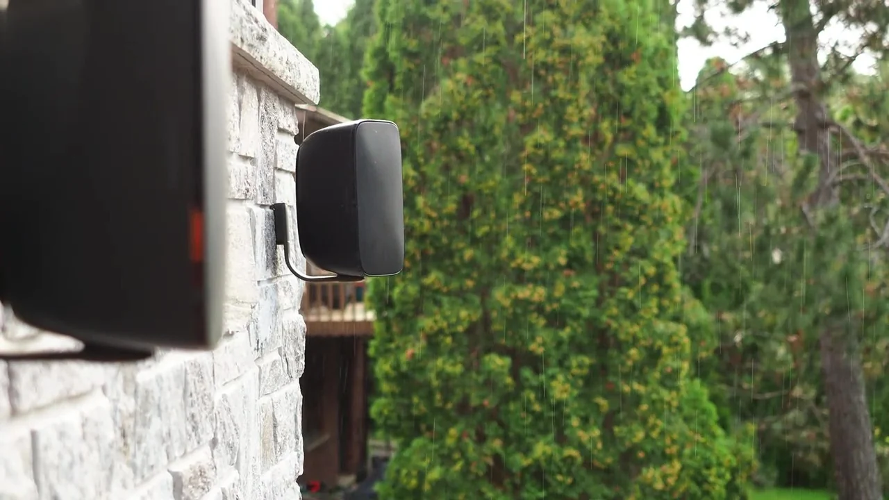 Bowers & Wilkins AM-1 outdoor speakers mounted on a stone wall beside a garden in the rain